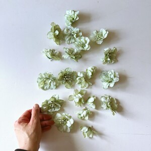 Può includere: Una collezione di fiori artificiali verde chiaro disposti su una superficie bianca. I fiori hanno più petali e un aspetto strutturato. Una mano si protende verso uno dei fiori.