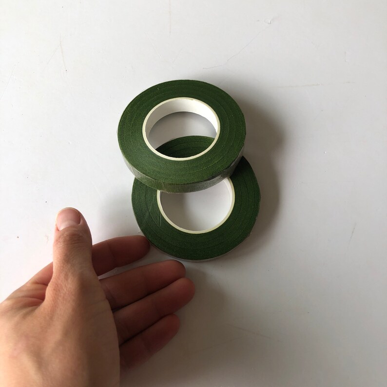 May include: Two rolls of dark green floral tape stacked on top of each other. The tape has a matte, textured surface and is wound around a white plastic core. The rolls are circular with a hollow center, showing the white inner ring.