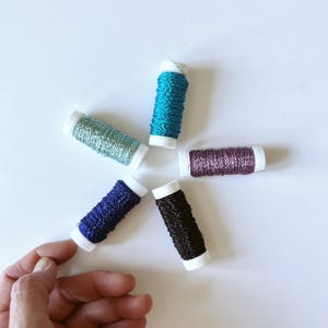Puede incluir: Cinco carretes de hilo de bordar metálico dispuestos en forma de estrella. Los colores incluyen turquesa brillante, verde menta claro, malva suave, azul real profundo y negro carbón oscuro. Cada carrete tiene extremos de plástico blanco y acabado brillante.