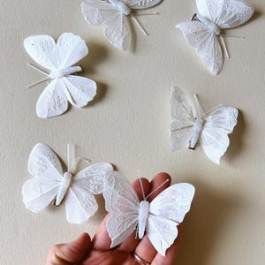 Op de afbeelding: Een set van vijf witte vlinderhaarclips met delicate, doorschijnende stoffen vleugels voorzien van ingewikkelde geborduurde patronen en dunne voelsprieten. Elke vlinder is bevestigd aan een kleine metalen clip met een lichtgewicht ontwerp.