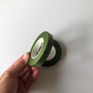 May include: Two rolls of dark green floral tape with a matte finish. Each roll features a central white paper core. The tape is designed for wrapping stems and is shown stacked slightly offset to reveal the circular shape and texture.