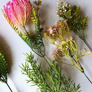 Puede incluir: Una colección de tallos botánicos artificiales que incluye una gran flor tipo protea rosa y naranja, un racimo de pequeñas flores rosas en ramas verdes delgadas, una ramita de bayas verdes y varios tallos de follaje verde texturizado.