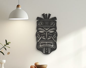 May include: Black metal tiki mask wall decor with intricate details and a fierce expression. The mask is mounted on a white wall above a wooden table.