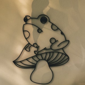 May include: A whimsical wall art piece featuring a stylized frog perched atop a mushroom. The design is rendered in a minimalist style with black outlines and circular details, creating a playful and decorative element. The artwork is suitable for a nursery or a kid's room.