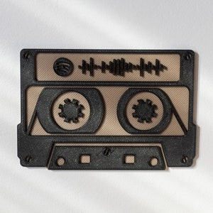 Op de afbeelding: Een zwart cassettebandontwerp met een beige midden en een geluidsgolfafbeelding. De cassetteband heeft twee cirkelvormige spoelontwerpen en een Spotify-logo. Het ontwerp staat op een witte achtergrond.
