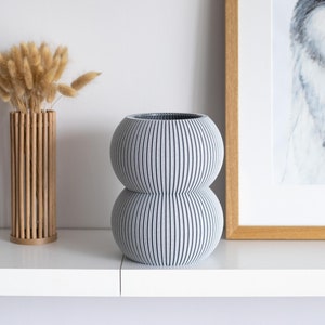 May include: A gray ceramic vase with a ribbed design. The vase has two sections, one on top of the other. The vase is sitting on a white shelf.