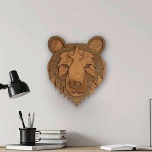 Puede incluir: Una decoración de pared de cabeza de oso de madera con un acabado de madera marrón. La cabeza de oso está montada en una pared blanca sobre un escritorio con una lámpara, lápices, libros y una imagen enmarcada.