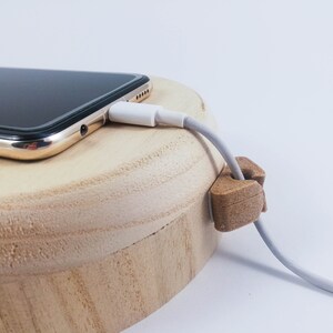 May include: A white charging cable is plugged into a gold smartphone resting on a wooden charging dock. A wooden cable organizer is holding the cable in place.