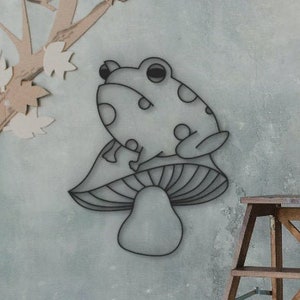 May include: A whimsical wall art piece featuring a frog perched atop a mushroom. The frog has a spotted pattern. The artwork is set against a muted blue-gray background, with a wooden ladder and a tree cutout in the scene.