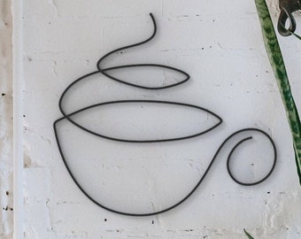 May include: A black metal wall art piece in the shape of a coffee cup with steam, fixed to a white brick wall. A potted plant with green leaves is positioned to the right of the artwork, adding a natural element.