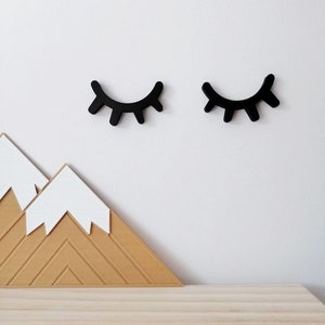 Könnte beinhalten: Schwarze Augenformen aus Holz mit Wimpern, montiert an einer weißen Wand über einem Holz-Wanddekor in Form eines Berges mit weißen Schneegipfeln.