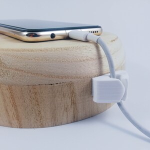 May include: A white cable management clip attached to a wooden surface, holding a white charging cable. A gold smartphone is resting on the wooden surface, with the charging cable plugged into the phone.