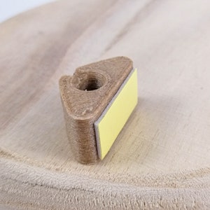 May include: A brown 3D printed object with a yellow adhesive backing. The object has a hole in the center and a triangular shape.