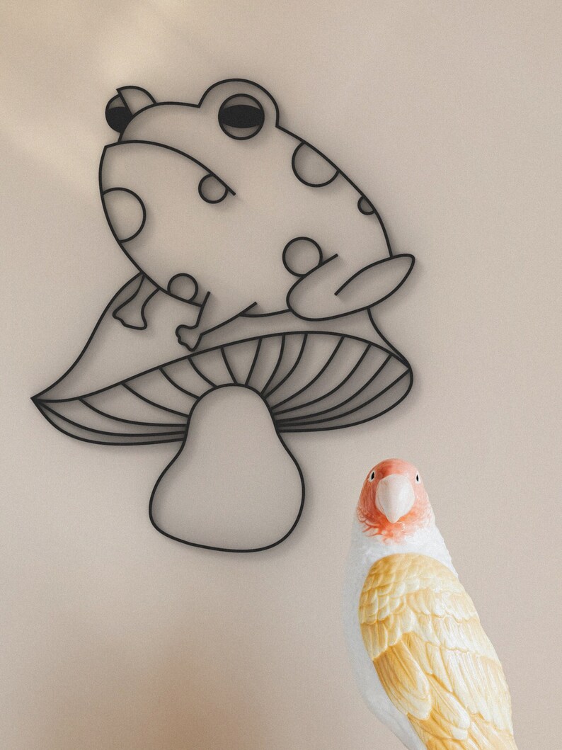 May include: Black metal frog wall art with white polka dots, perched on a mushroom. A decorative bird with yellow and white feathers and a red beak is in the foreground. The art is set against a neutral background.