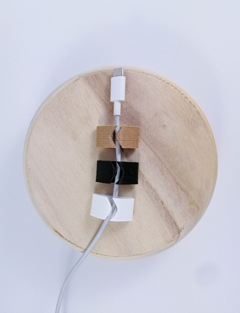 May include: A white cable is held in place by three cable clips. The clips are made of wood, black plastic, and white plastic. The cable is on a wooden surface.