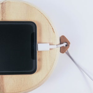 May include: A black smartphone is charging on a wooden charging dock. A white charging cable is connected to the phone and secured with a brown cable organizer.