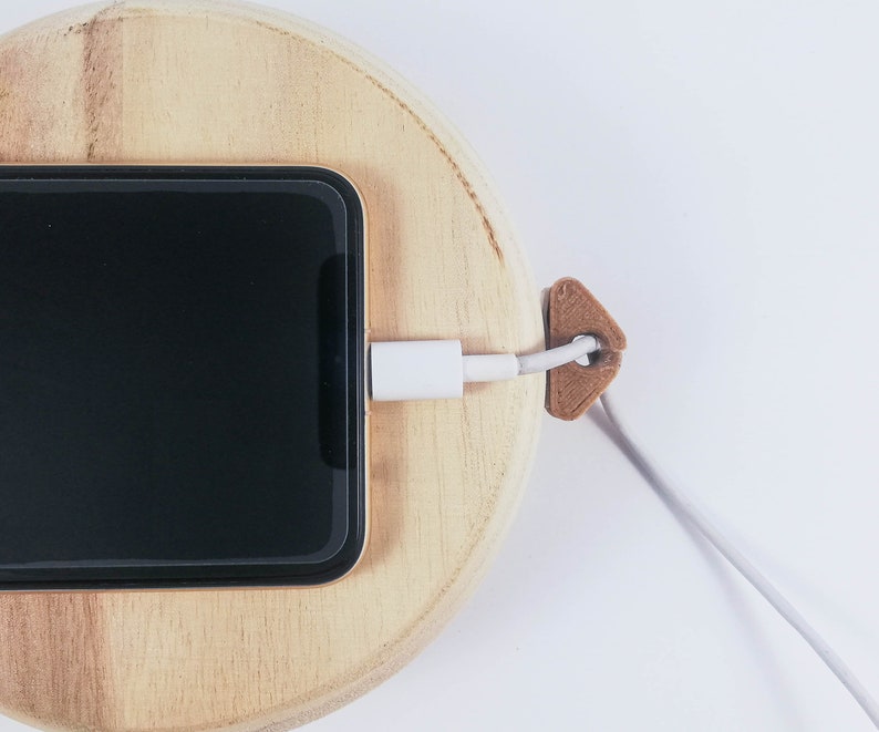 May include: A black smartphone is charging on a wooden charging dock. A white charging cable is connected to the phone and secured with a brown cable organizer.