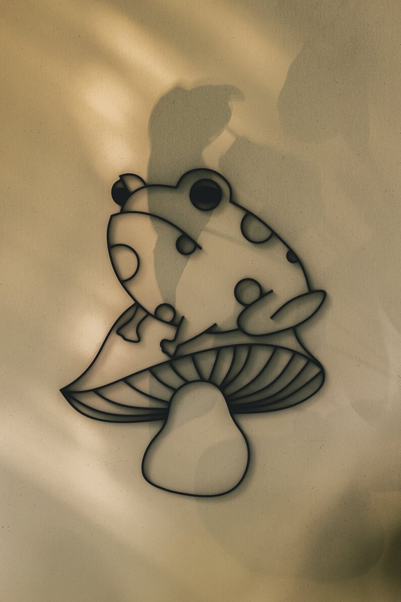 May include: A whimsical wall art piece featuring a stylized frog perched atop a mushroom. The design is rendered in a minimalist style with black outlines and circular details, creating a playful and decorative element. The artwork is suitable for a nursery or a kid's room.
