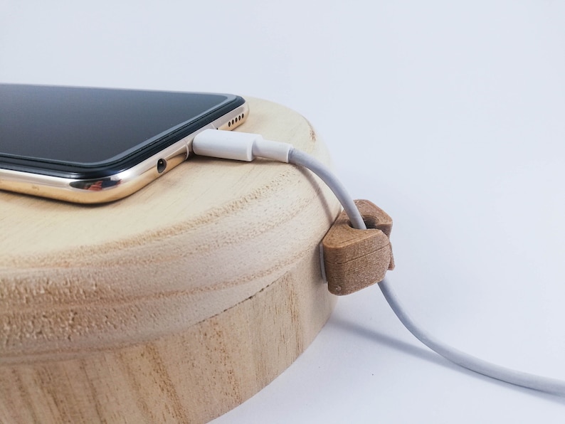 May include: A white charging cable is plugged into a gold smartphone resting on a wooden charging dock. A wooden cable organizer is holding the cable in place.