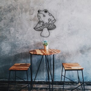 May include: A whimsical wall art piece featuring a frog perched atop a mushroom. Below, a small round wooden table with black metal legs and two matching stools. A small vase of flowers sits on the table. The background is a textured gray wall.
