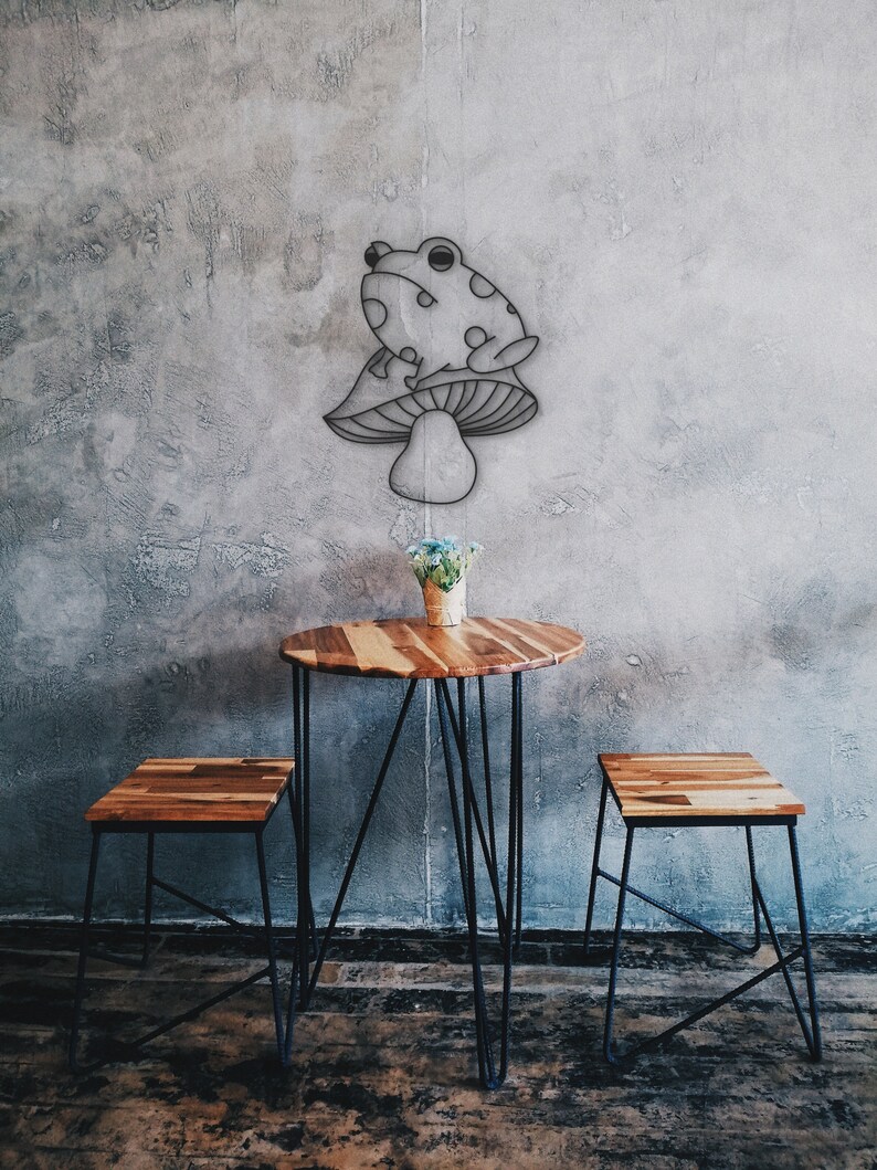 May include: A whimsical wall art piece featuring a frog perched atop a mushroom. Below, a small round wooden table with black metal legs and two matching stools. A small vase of flowers sits on the table. The background is a textured gray wall.