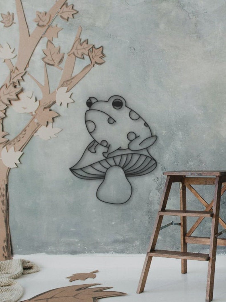 May include: A whimsical wall art piece featuring a frog perched atop a mushroom. The frog has a spotted pattern. The artwork is set against a muted blue-gray background, with a wooden ladder and a tree cutout in the scene.