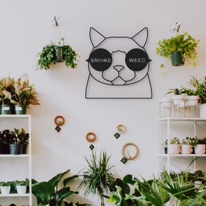 Pode incluir: Arte de parede de metal preto com um gato usando &oacute;culos de sol com as palavras "SMOKE" e "WEED" impressas nas lentes. O gato est&aacute; rodeado de plantas em vasos.