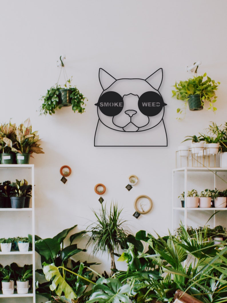 Pode incluir: Arte de parede de metal preto com um gato usando &oacute;culos de sol com as palavras "SMOKE" e "WEED" impressas nas lentes. O gato est&aacute; rodeado de plantas em vasos.