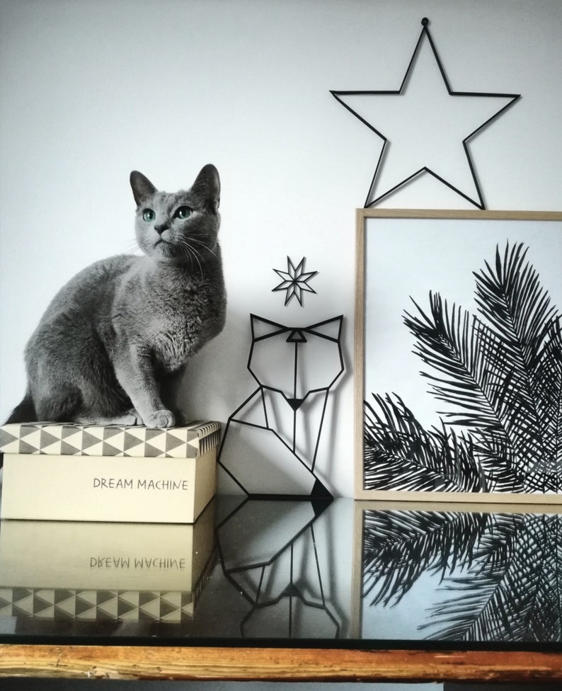May include: A gray Russian Blue cat sits on a box with the text "DREAM MACHINE" printed on it. The cat is looking to the left of the image. Behind the cat is a black metal geometric fox sculpture and a black metal star hanging on the wall. A framed print with a black and white image of palm leaves is on the wall to the right of the cat.