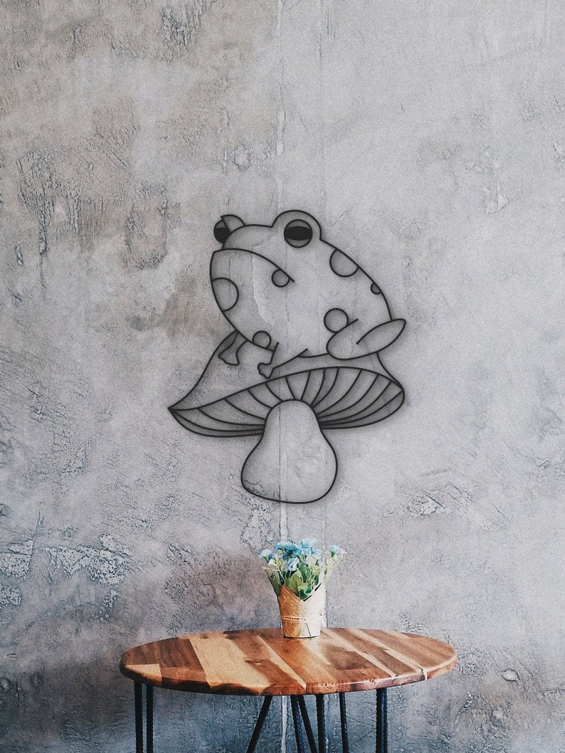 May include: A metal wall art piece featuring a frog sitting on a mushroom. The frog has large eyes and polka dots. The mushroom has a rounded cap and stem. The art is mounted on a textured gray wall.