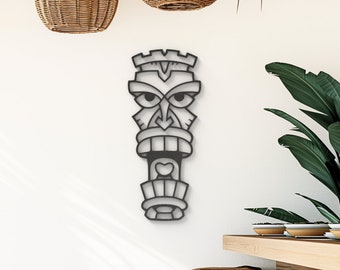 May include: Black metal tiki mask wall decor with a stylized face and geometric patterns. The mask is mounted on a white wall with a wooden table and chairs in the background.