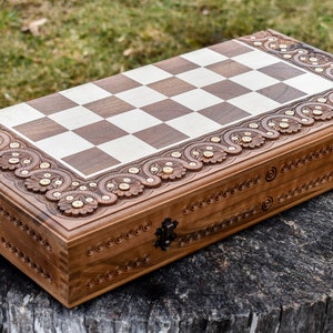 Op de afbeelding: Een houten schaakbord met ingewikkelde houtsnijwerk rond de rand. Het bord is gesloten en heeft een donkerbruine afwerking. Het schaakbord staat op een boomstronk.