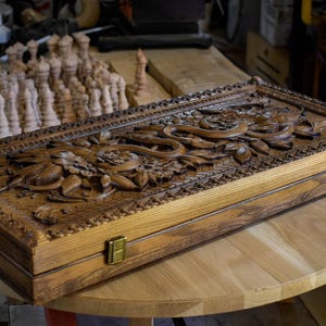 May include: A rectangular wooden backgammon set with intricate floral and serpent carvings on the lid. The set is made of light brown wood, with brass-colored clasps. Chess pieces are visible in the background.