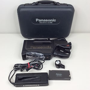 Puede incluir: Videocámara VHS Panasonic OmniMovie negra con accesorios, que incluyen una funda de transporte, un cargador de batería, una batería y una luz. La videocámara tiene el logotipo de Panasonic y "x8" en el lateral. Los accesorios están colocados alrededor de la videocámara.