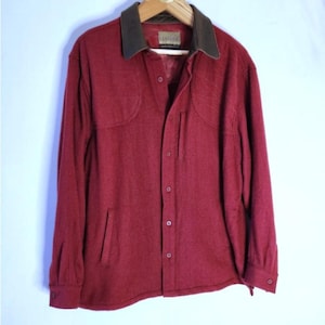 May include: A long-sleeved button-down shirt in a deep maroon color featuring a contrasting dark brown leather collar. The shirt includes a vertical zippered chest pocket, two lower side pockets, and buttoned cuffs, hanging on a wooden hanger.