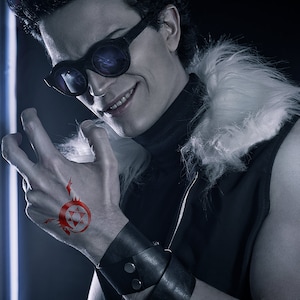 May include: A man with dark hair and a smirk is wearing a black vest with a white fur collar. He has a red tattoo on his hand of a circle with a star inside. He is wearing black sunglasses and a black leather bracelet.
