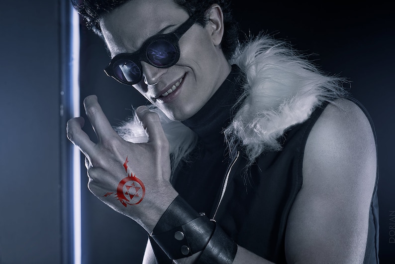 May include: A man with dark hair and a smirk is wearing a black vest with a white fur collar. He has a red tattoo on his hand of a circle with a star inside. He is wearing black sunglasses and a black leather bracelet.
