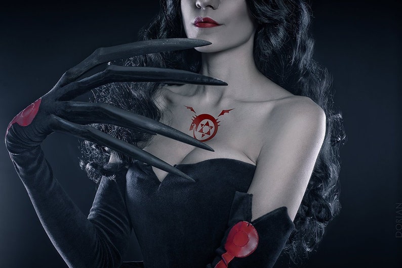 May include: A woman with long black hair and red lipstick wears a black strapless dress and black gloves with long sharp claws. A red symbol with a star and a snake is on her chest.