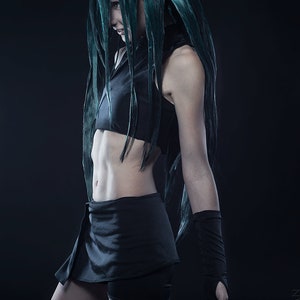 May include: A woman with long, dark green hair is wearing a black crop top and a short black skirt. She has a red tattoo on her thigh that features a star and a circle with a lightning bolt.