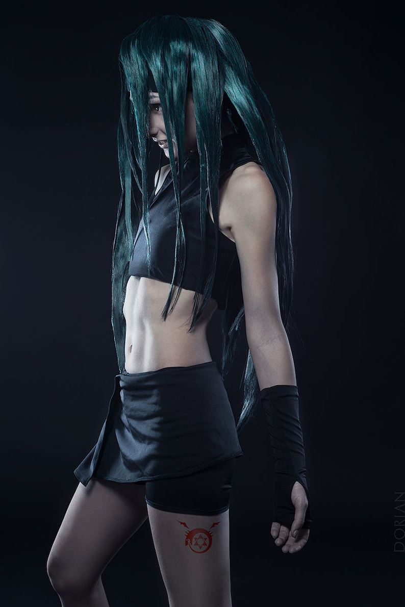 May include: A woman with long, dark green hair is wearing a black crop top and a short black skirt. She has a red tattoo on her thigh that features a star and a circle with a lightning bolt.