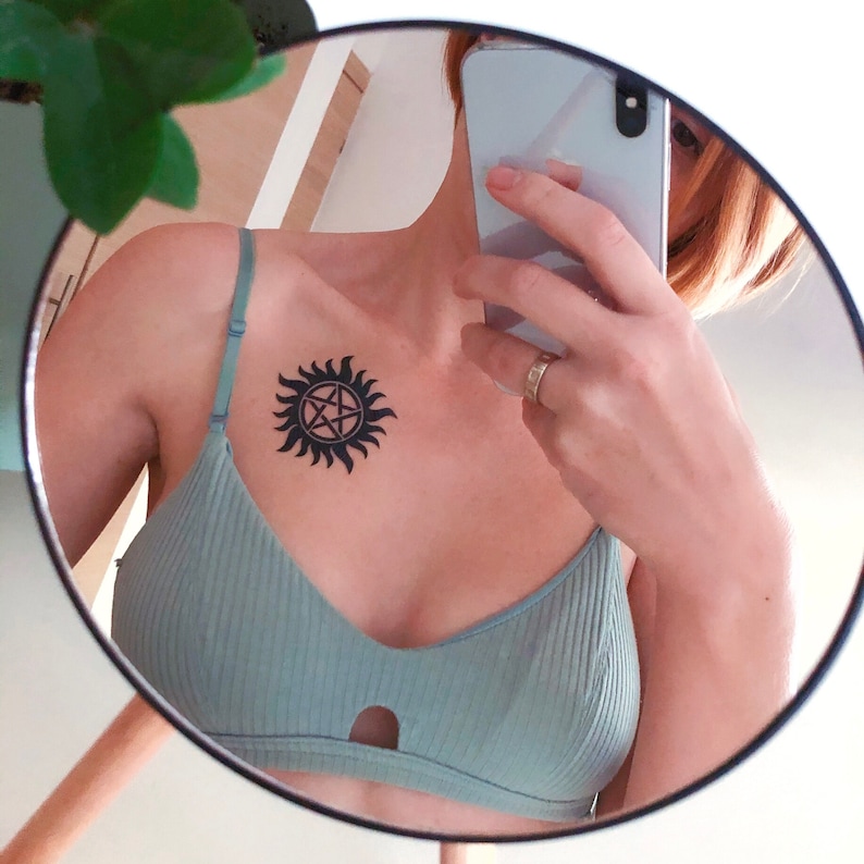 May include: A close-up of a person's chest with a black and white tattoo of a pentagram inside a circle. The tattoo is on the left side of the chest, just above the cleavage. The person is wearing a light green bra with a keyhole opening in the front.