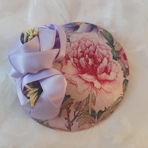 Könnte beinhalten: Ein kleiner, runder Hut mit einem Blumendruck in Rosa-, Grün- und Brauntönen. Eine große lila Blume mit gelben Akzenten ist am Hut befestigt.