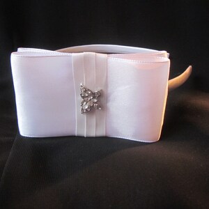 May include: A white satin ribbon headband featuring a large, structured bow at the centre. The bow is accented with a silver-toned butterfly-shaped embellishment encrusted with small, clear, reflective stones positioned on a vertical ribbon band.
