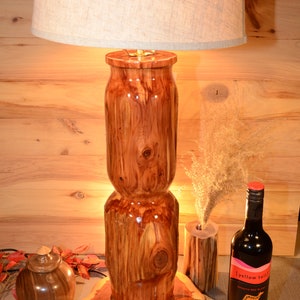 Puede incluir: Una lámpara de madera con un acabado de madera natural y una pantalla blanca. La base de la lámpara está hecha de una sola pieza de madera y tiene una forma única y orgánica. La lámpara está sobre una mesa de madera junto a una botella de vino.