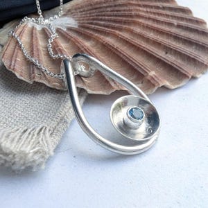 May include: A silver pendant necklace with a spiral design and a small, round, blue gemstone at the center. The pendant hangs from a delicate silver chain. The necklace is displayed on a seashell.