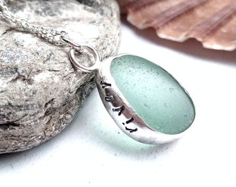 May include: A silver necklace with a light green sea glass pendant. The pendant is set in silver and has the words "It's a 11" engraved on it.