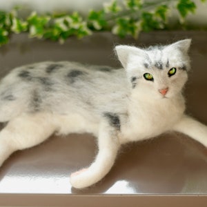 May include: A white and gray felted cat with green eyes lying on a brown surface.