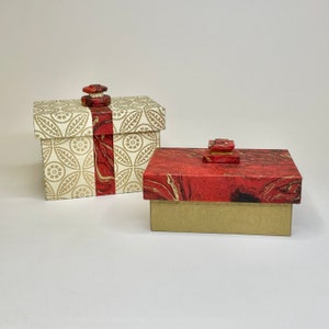 May include: Two decorative boxes with red and gold marbled paper designs. The larger box has a white background with gold floral patterns and a red ribbon. The smaller box has a red background with gold marbling.
