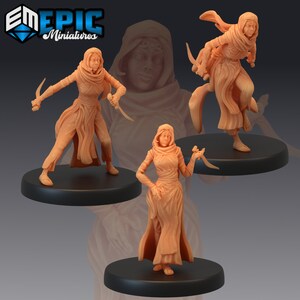 May include: Three miniature figures of women wearing hooded robes and holding daggers. The figures are painted in a light brown colour and are standing on black bases.