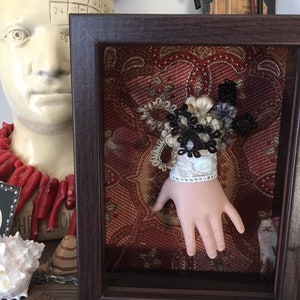 Pode incluir: Uma obra de arte emoldurada com uma mão de boneca adornada com uma pulseira de renda e um buquê de elementos decorativos pretos, brancos e bege. O fundo é um tecido vermelho estampado. A moldura é marrom escuro.